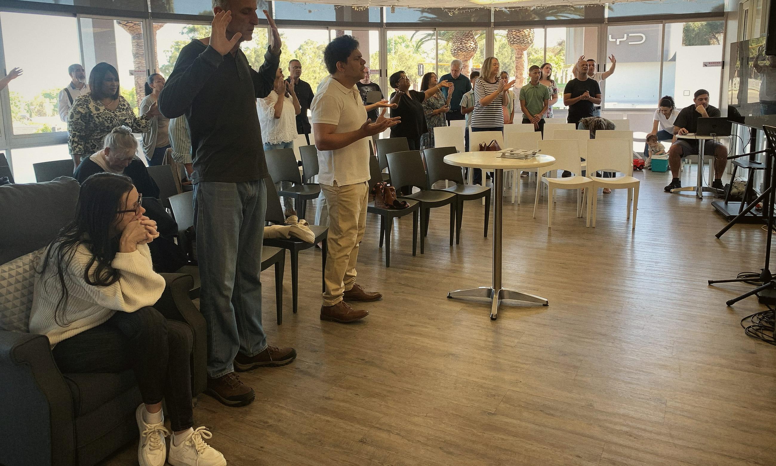 The congregation standing to worship in a bright room, hands raised, palm trees through the windows behind them.