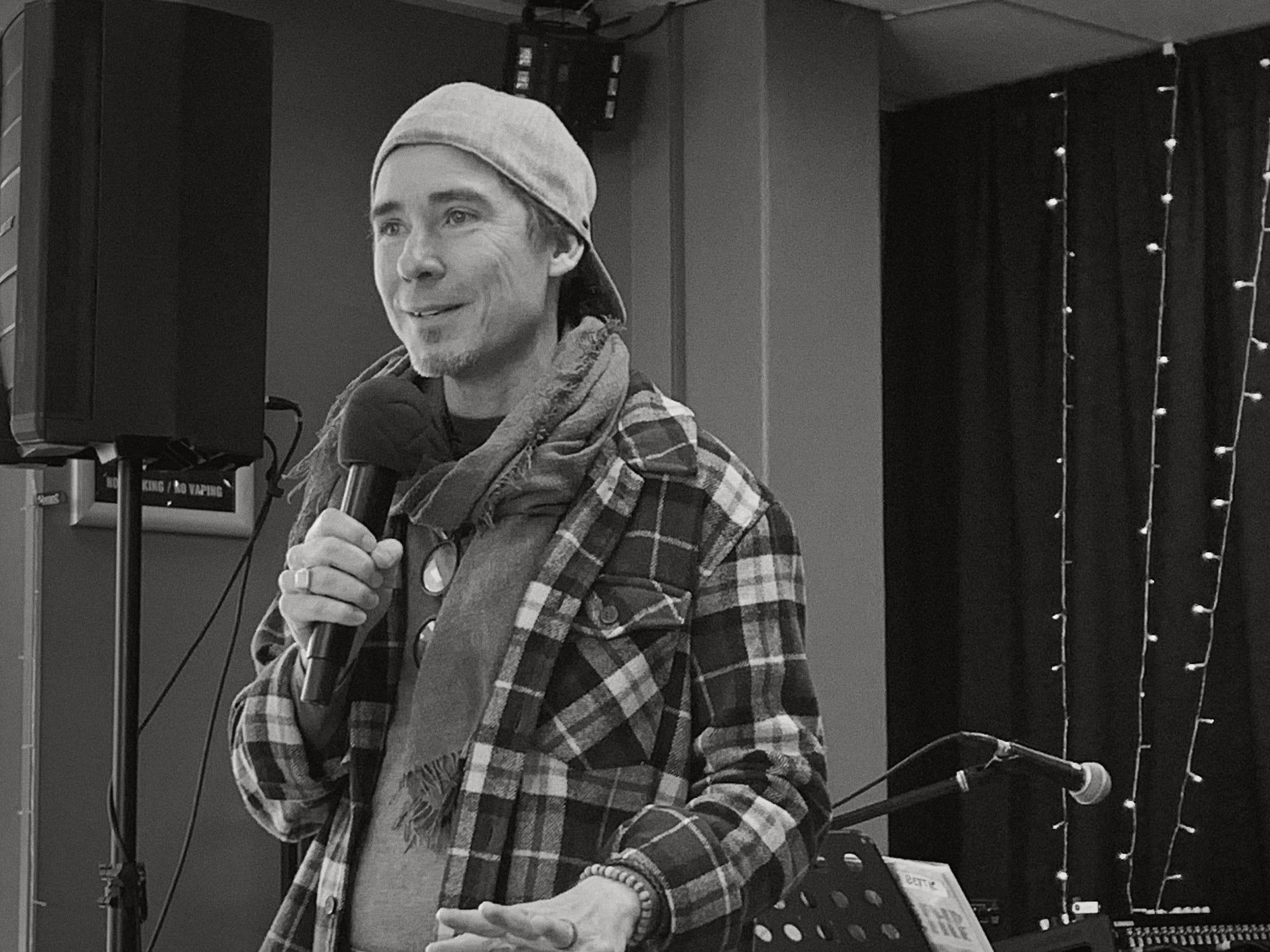 A man in a flannel shirt and cap speaking into a microphone, smiling, fairy lights behind him.